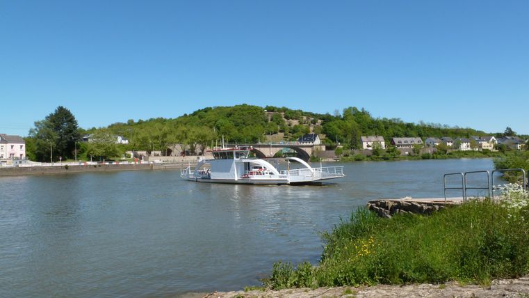 A ship sails on a calm river, surrounded by green hills. The sky is clear and blue.
