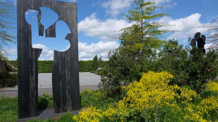 Eine moderne Skulptur steht zwischen bunten Blumen und grünem Strauch. Der Himmel ist klar mit einigen Wolken.