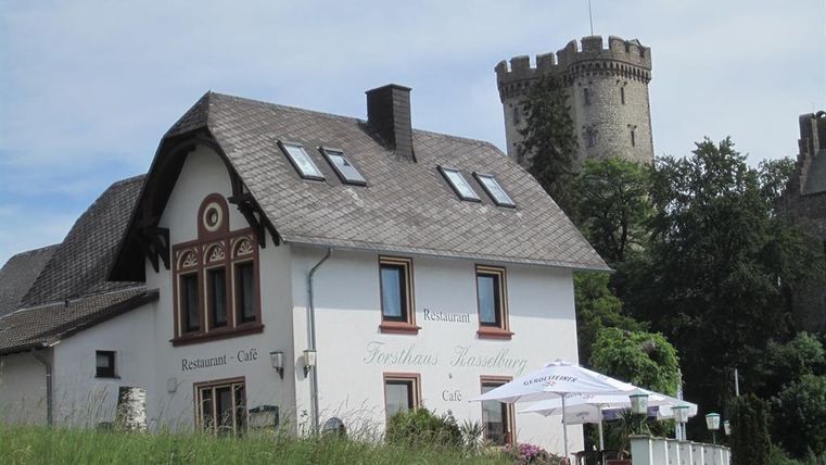 White building of the Kasselburg forestry house with café and restaurant, in the background a stone tower with a German flag.