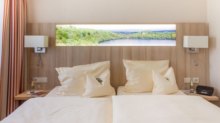 Ein gemütliches Doppelbett mit weißen Bettdecken und Kissen. Im Hintergrund ist ein Fenster mit Blick auf eine grüne Landschaft und einen Gewässer.