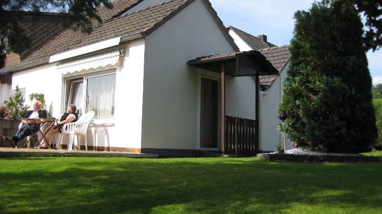 Ein helles Haus mit einem gepflegten Garten. Auf der Terrasse sitzen Personen und genießen die Sonne.