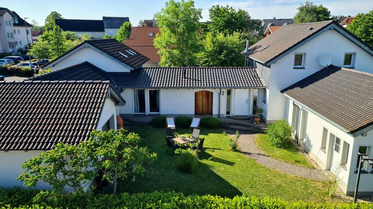 Ein Garten mit zwei Liegestühlen und einem Weg, umgeben von grünen Pflanzen. Im Hintergrund sind mehrere Häuser sichtbar, unter blauem Himmel.