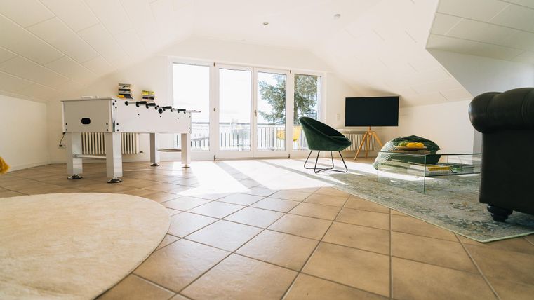 Een lichte woonkamer met een tafelvoetbalspel, een comfortabele fauteuil en een televisie. Grote ramen zorgen voor veel natuurlijk licht.