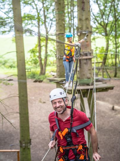 Ein Mann mit einem Helm und Sicherheitsgurt lächelt, während er an Seilen zwischen Bäumen schwingt. Im Hintergrund steht eine weitere Person auf einer Plattform.