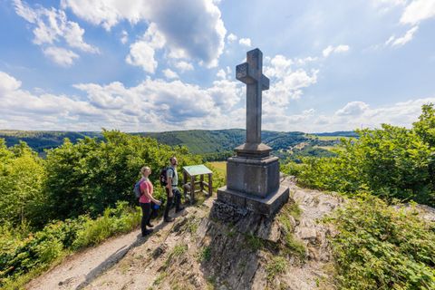 A group of people stands by a cross on a hill overlooking the green landscape. The sky is clear with some clouds.