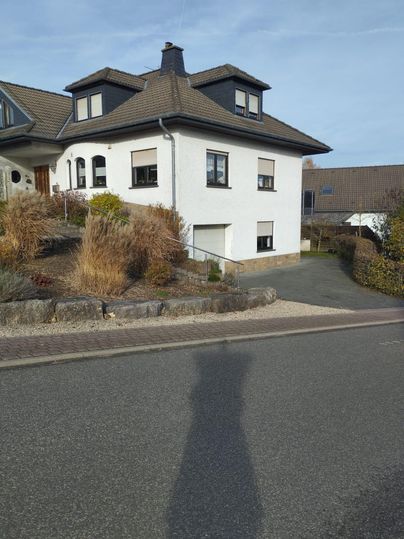 A modern two-story house with a landscaped front garden. The facade is white and the roof is dark.