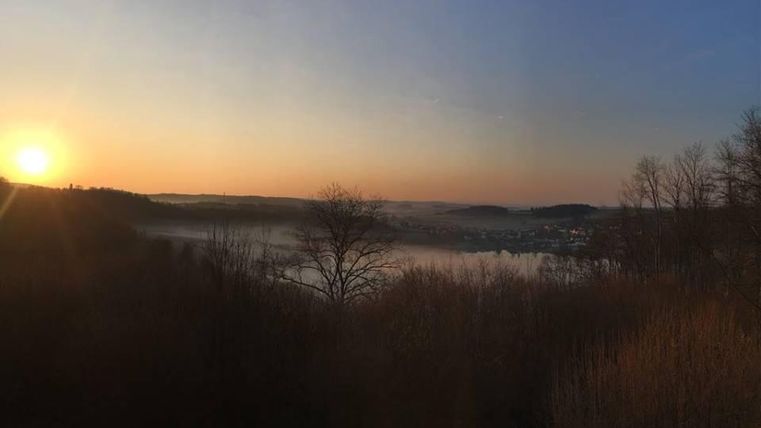 A picturesque landscape at sunrise with fog over the water. The trees in the foreground create a serene atmosphere.
