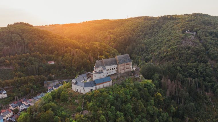 Luftaufnahme der Burg Vianden in Luxemburg bei Sonnenuntergang, umgeben von bewaldeten Hügeln.