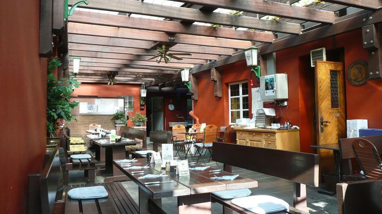 Ein einladender Außenbereich eines Restaurants mit Holzmöbeln und begrünten Wänden. Die Sonneneinstrahlung sorgt für eine freundliche Atmosphäre.