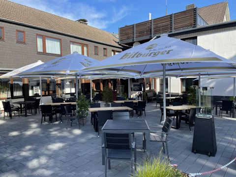 Een uitnodigend terras met tafels en stoelen onder grote parasols. Op de achtergrond is een gebouw te zien dat een ontspannen sfeer creëert.