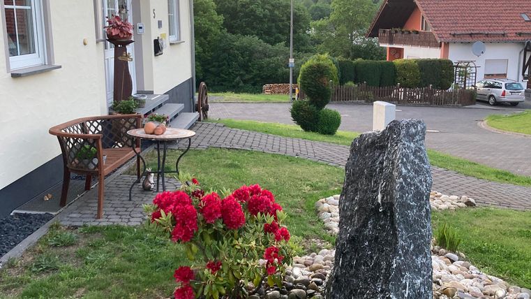 Een gezellige hoek van het huis met een bloementuin en een decoratieve steen. Op de achtergrond zijn heuvels en een heldere lucht te zien.