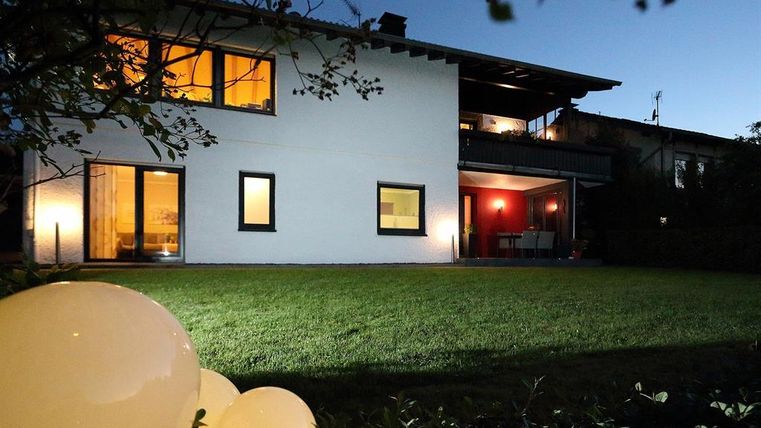 Een modern huis met grote ramen, verlicht 's nachts. De tuin is groen en heeft decoratieve verlichtings elementen.