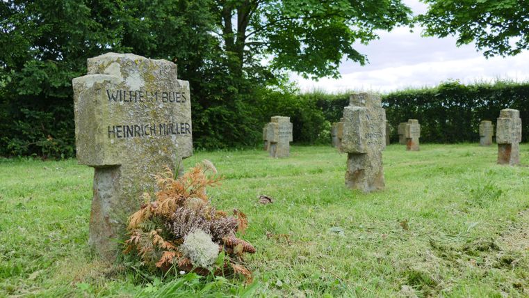 Ein Friedhof mit mehreren Grabsteinen aus Stein, auf einer grünen Wiese. Einige Grabsteine haben Namen eingraviert und es liegt ein kleiner Blumenstrauß auf einem der Gräber.