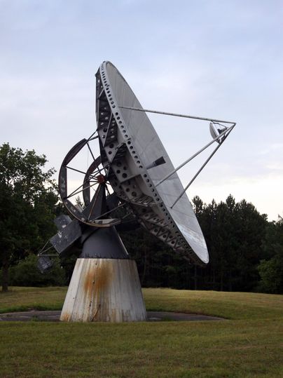 Eine große Satellitenantenne steht auf einem Rasen. Im Hintergrund sind Bäume und ein bewölkter Himmel sichtbar.