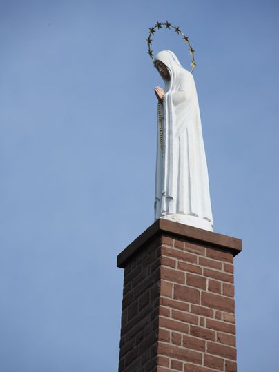Eine weiße Marienstatue mit Heiligenschein steht auf einer Backsteinsäule vor blauem Himmel.