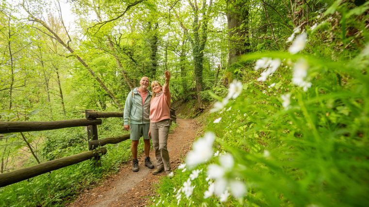 Een stel staat glimlachend op een wandelpad in een groene bos. Om hen heen bloeien witte bloemen en de sfeer is fris en uitnodigend.