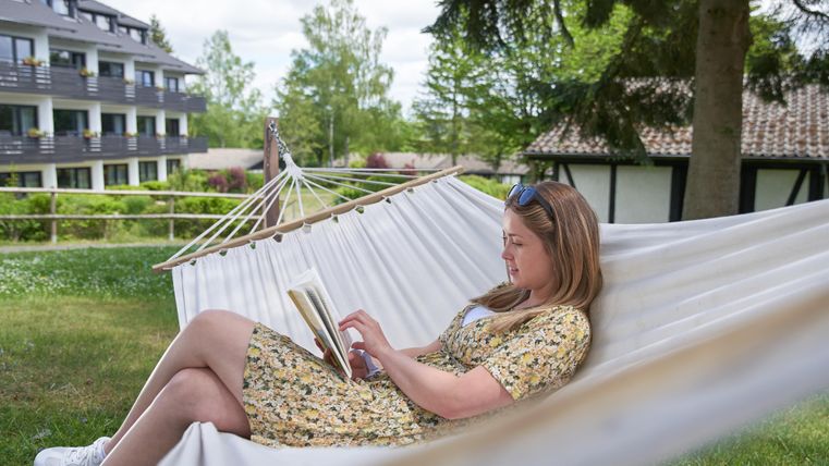 Eine entspannte Frau sitzt in einer Hängematte und liest ein Buch. Im Hintergrund sind grüne Bäume und moderne Gebäude zu sehen.