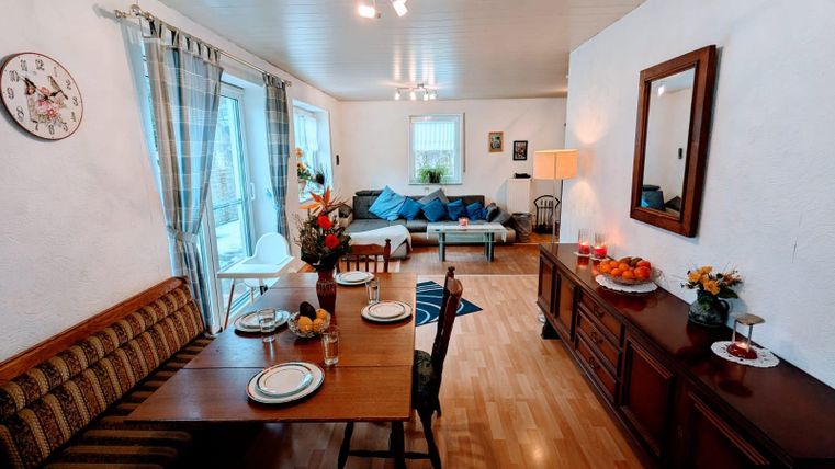 A cozy living room with a dining area and a sofa. The room is bright and inviting, with flowers and decorative elements.