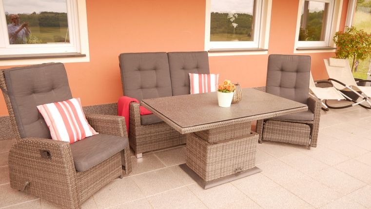 A cozy terrace with rattan-like furniture and comfortable cushions. The table is in the center and a flower pot adds to the inviting atmosphere.