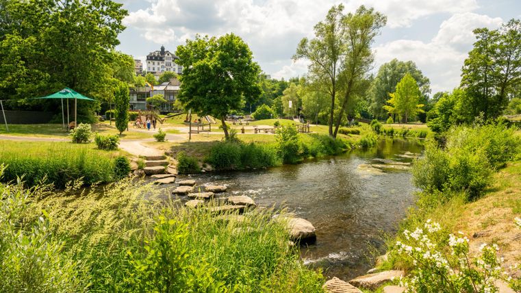 Een idyllisch park met een rivier, bomen en een speeltuin. Mensen wandelen en genieten van de natuur. Op de achtergrond een groot gebouw.