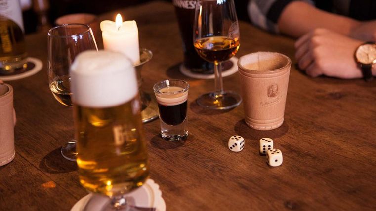 Ein Tisch mit verschiedenen Getränken, darunter Bier, Schnaps und ein Glas Whisky. Daneben liegen zwei Würfel und Kerzenlicht für eine gemütliche Atmosphäre.