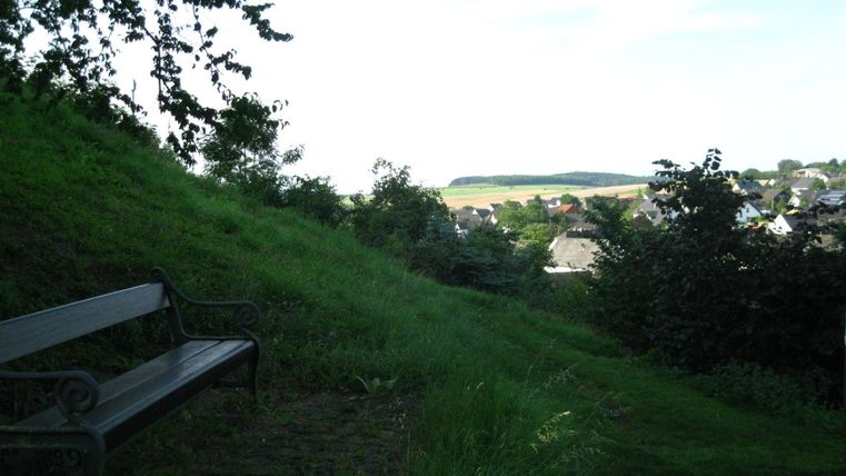 A quiet landscape with a bench in the foreground and green meadows in the background. In the background, some houses and a gentle hill can be seen.