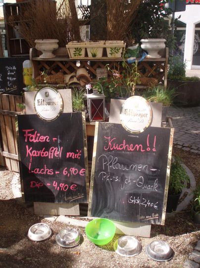 Ein gemütlicher Außenbereich mit zwei Tafeln, die Gerichte und Preise anzeigen. Auf den Tafeln stehen die Angebote von Fischgerichten und Kuchen.