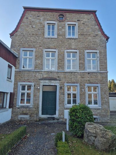 A historic stone house with a green front door and large windows. Surrounded by a small garden and a few stones in the foreground.