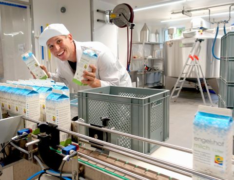 Ein fröhlicher Mitarbeiter in einer Produktionsstätte hält Milchkartons in der Hand. Im Hintergrund sind Maschinen und Ausrüstungen zu sehen.