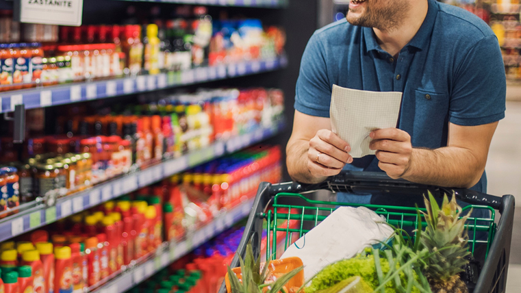Ein Mann mit einem gut gefüllten Einkaufswagen läuft durch einen Gang im Supermarkt. In den regalen sind Soßen und Ketchup zu sehen. In seiner Hand hält er eine Einkaufsliste