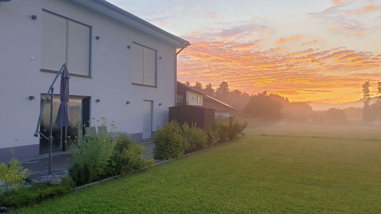 Ein modernes Haus am Morgen mit einem wunderschönen Sonnenaufgang im Hintergrund. Der Garten ist grün und gut gepflegt, umgeben von leichtem Nebel.