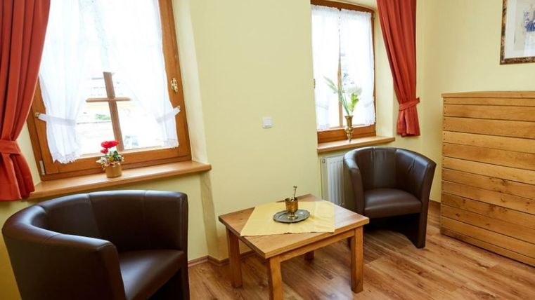 Ein gemütlicher Raum mit zwei schwarzen Sesseln und einem kleinen Holztisch. Die Wände sind gelb, und es gibt große Fenster mit roten Vorhängen.