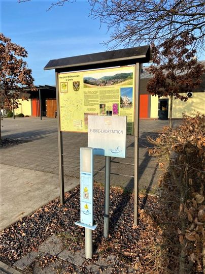 Ein Informationsschild über die Umgebung und eine E-Bike-Ladestation. Im Hintergrund sind Gebäude und strahlend blauer Himmel zu sehen.