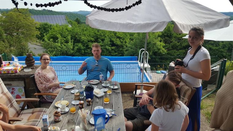 Ein geselliger Frühstückstisch im Freien mit mehreren Personen. Im Hintergrund ist ein Pool und viel grüne Landschaft zu sehen.