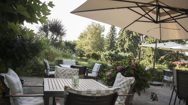 Een gezellige zithoek in de tuin met tafels en stoelen onder parasols. Omgeven door een groene omgeving en bloeiende planten.