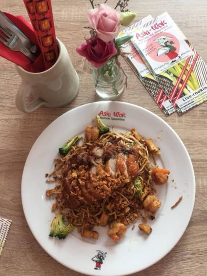 Een bord met gebakken noedels en verschillende ingrediënten zoals groenten en garnalen. Op de tafel staat een vaasje met een bloem en Chinese bestek.
