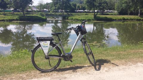 A bike tour in nature with an e-bike, standing by a quiet lake. In the background, there are trees and a campsite.
