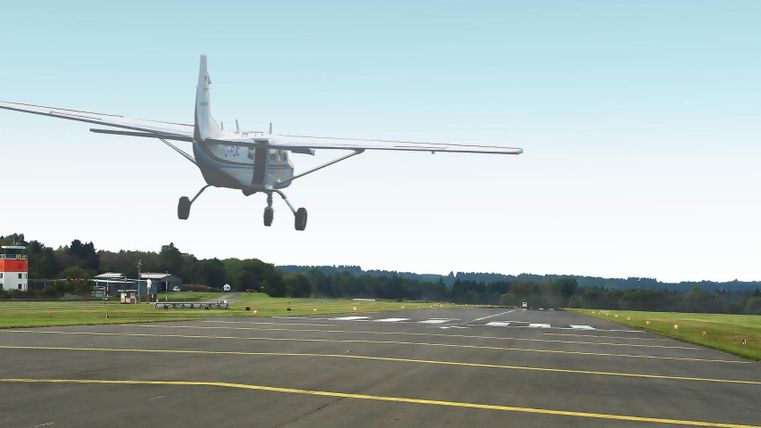 Een klein vliegtuig landt op een luchthaven. Op de achtergrond zijn de groene velden en een gebouw te zien.