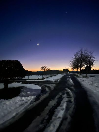 A snowy landscape at dusk with a winding path. In the sky, two luminous objects are visible above a gentle gradient.