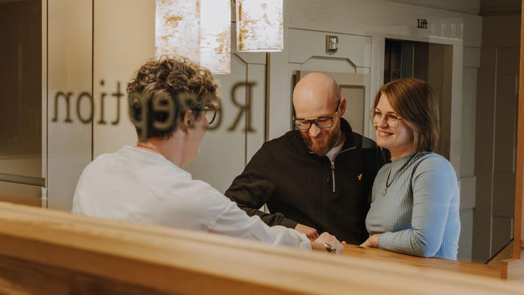 Ein Paar steht am Empfangstresen und spricht mit einer Mitarbeiterin. Im Hintergrund sind dekorative Lampen und ein Spiegel zu sehen.