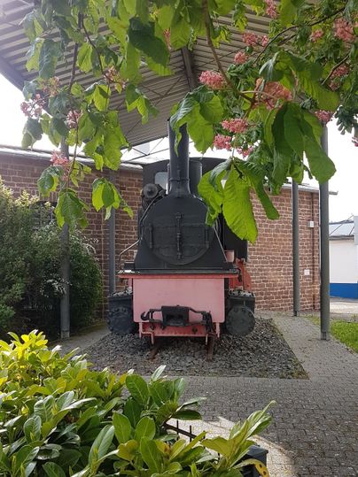 Eine alte Dampflok steht unter einem Vordach, umgeben von grünen Pflanzen und blühenden Bäumen. Im Hintergrund sieht man eine Ziegelmauer und moderne Gebäude.