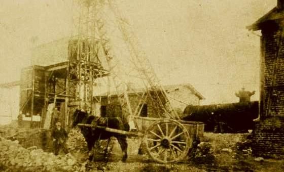 An old image of a construction site with a steam locomotive and a hoist structure. A horse is pulling a cartload near the workplace.