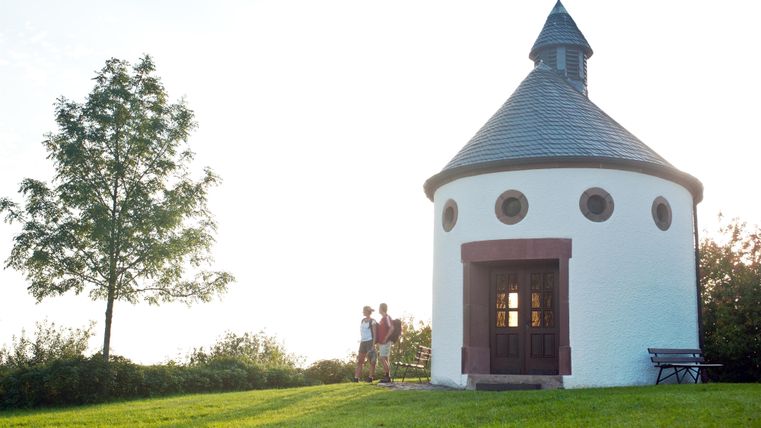 Runde Kapelle mit Spitzdach und Kreuz, zwei Personen stehen daneben. Ein Baum und eine Bank auf einer grünen Wiese.