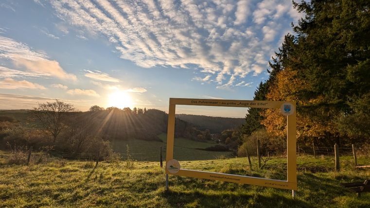 Een schilderachtig uitzicht op een groene landschap met een zonsondergang op de achtergrond. Op de voorgrond staat een gele lijst die als fotopunt dient.