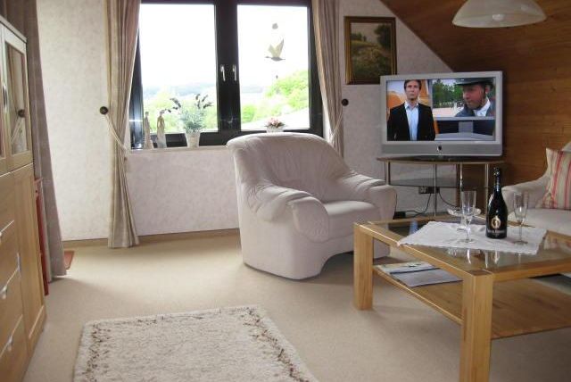 A cozy living room with a white armchair and a wooden table. In the background, a television is showing a program.