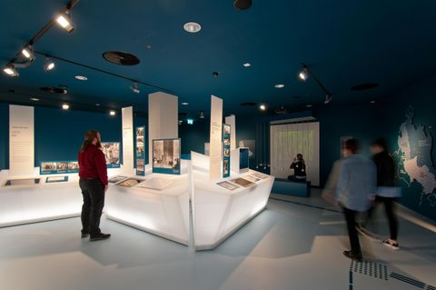 Ein modernes Ausstellungsraum mit blauen Wänden und hellem Display. Besucher interagieren mit verschiedenen Exponaten in der Mitte des Raumes.