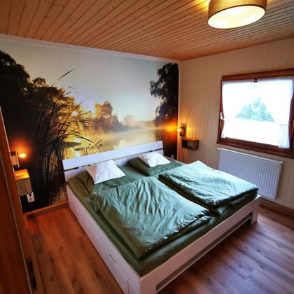 Een gezellige slaapkamer met een groot bed en groene beddengoed. Aan de muur is een landschappelijk schilderij te zien met een meer en bomen.