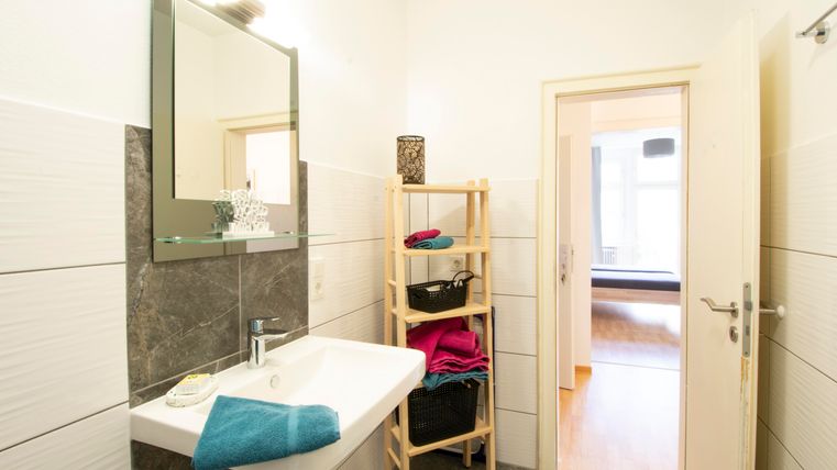 Ein modernes Badezimmer mit einem Waschbecken und einem Spiegel. Es gibt einen Regalschrank mit handtüchern und einen Blick auf einen weiteren Raum.