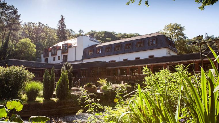 Ein malerisches Hotel umgeben von üppigem Grün und einem ruhigen Wasserlauf. Die Fassade des Gebäudes ist modern und einladend.