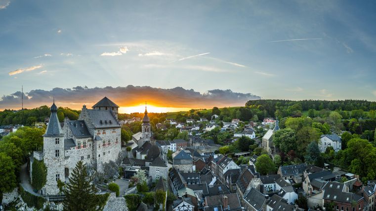 Eine beeindruckende Aussicht auf eine Burg bei Sonnenuntergang. Die umliegende Landschaft ist grün und die Gebäude der Stadt sind gut sichtbar.